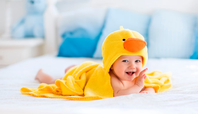 Foto: Fröhliches lachendes Baby mit Badetuch im Entendesign, das auf dem Bett der Eltern nach dem Bad oder der Dusche liegt.